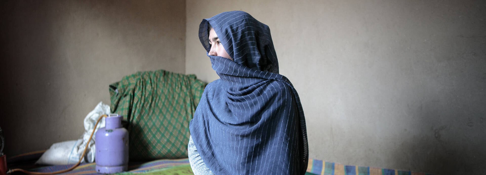 Eine Frau aus Afghanistan sitzt in ihrem Zuhause und schaut zur Seite. Ein blaues Tuch bedeckt ihren Kopf und ihren Mund.