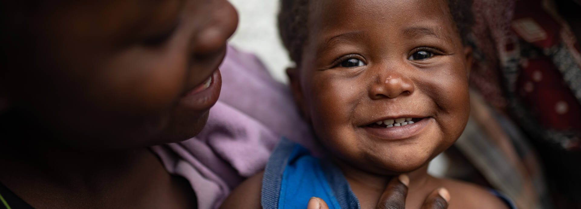 Ein Kleinkind schaut lachend in die Kamera, während die Mutter es beschützend hält und anlächelt