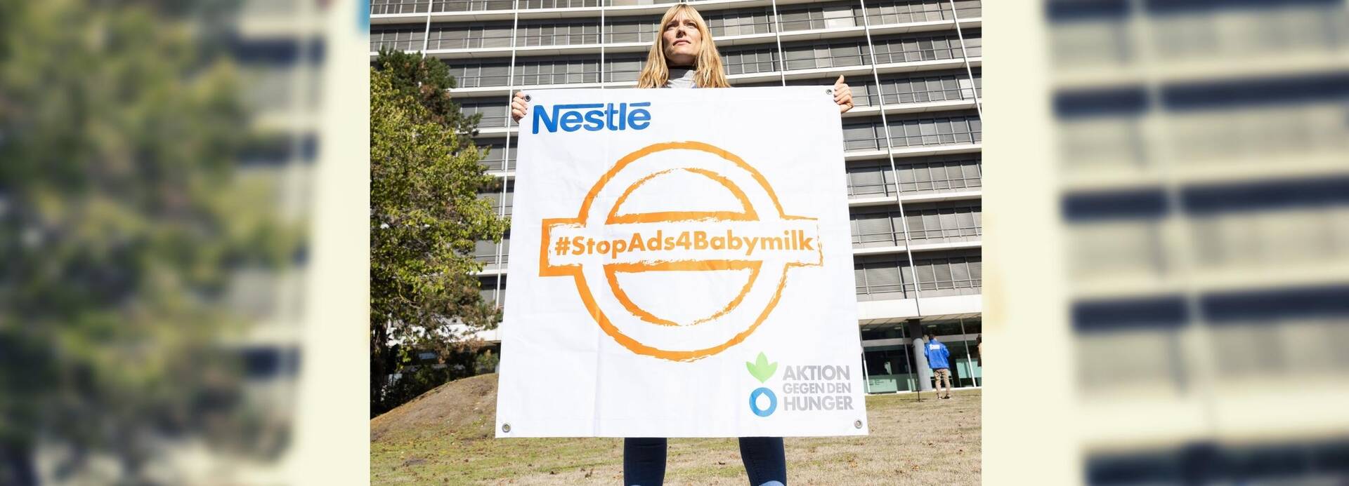 Unsere Kollegin steht vor der Nestlé-Zentrale in Frankfurt am Main und hält ein Banner mit einem orangefarbenen Stempel mit dem Hashtag #StopAds4Babymilk hoch.