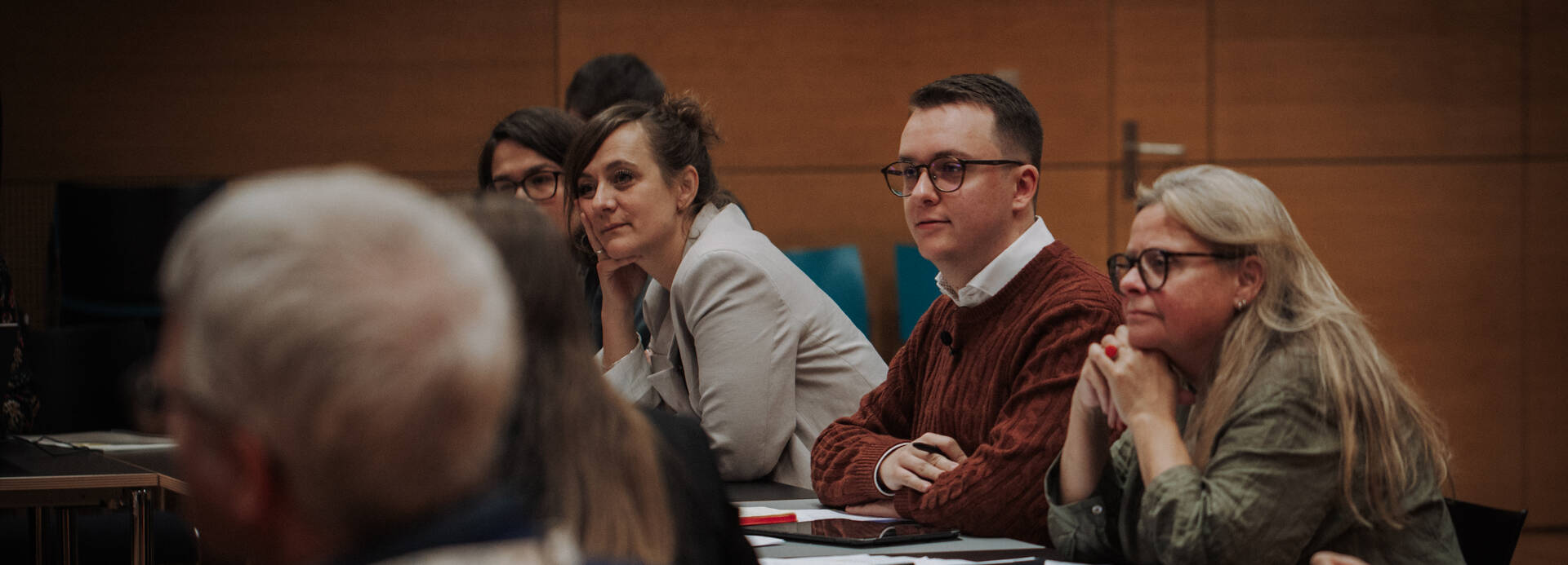 Der Moderator und zwei Teilnehmerinnen sitzen am Tisch und hören konzentriert zu.