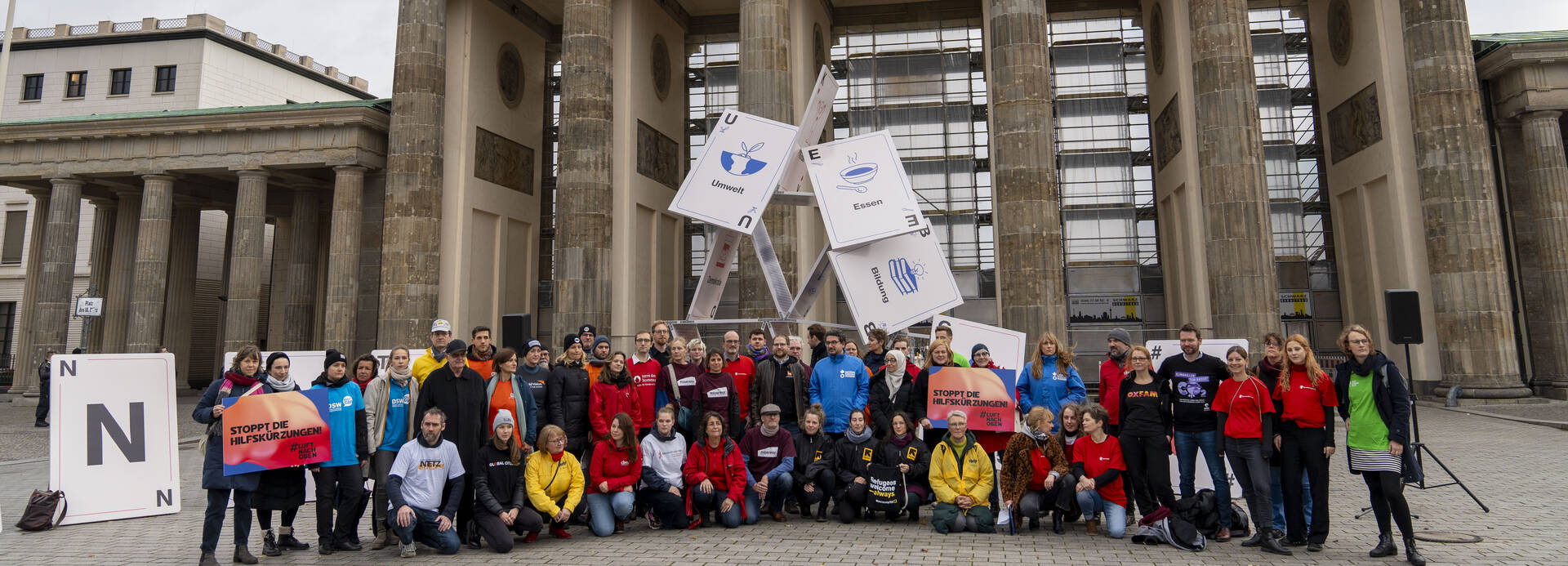 Mitglieder von Hilfsorganisationen bei der Protestaktion #Luftnachoben vor dem Brandenburger Tor in Berlin, im Hintergrund ein zusammenfallendes Kartenhaus.