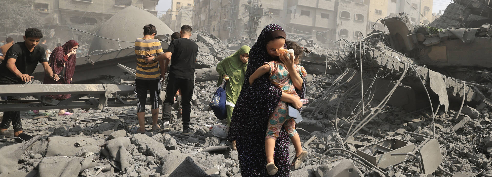 Eine Frau trägt ihr Kind durch eine von Angriffen zerstörte Straße in Gaza Stadt. Eine Frau trägt ihr Kind durch eine von Angriffen zerstörte Straße in Gaza Stadt, im Hintergrund weitere Menschen.