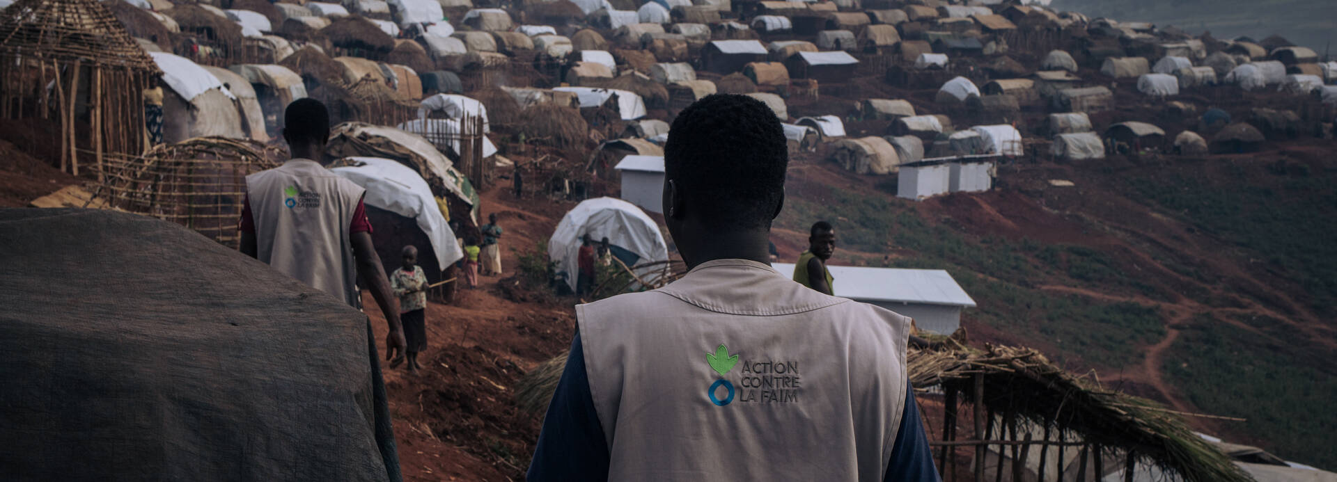 Zwei Mitarbeiter von Aktion gegen den Hunger von hinten in der Nähe eines Geflüchtetencamps in der Demokratischen Republik Kongo, auf den Westen das Logo