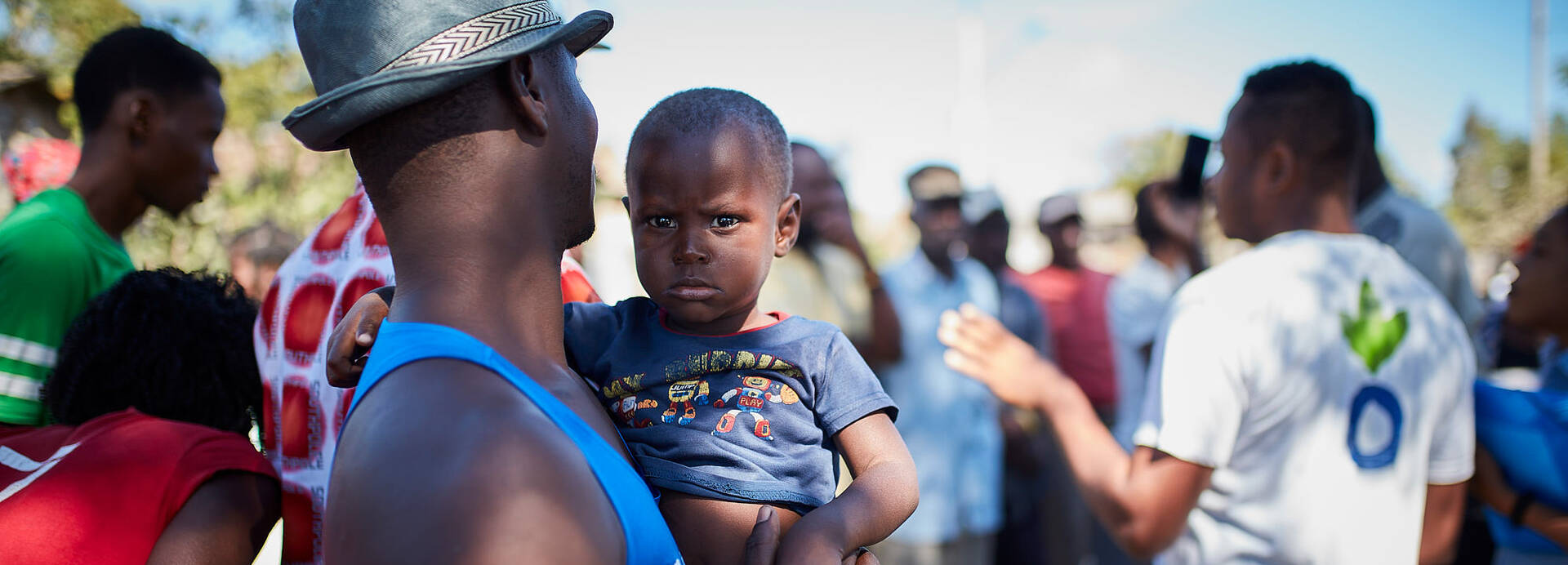 Ein Mann mit Hut trägt ein Kind auf dem Arm, während er einer Erklärung eines unserer Mitarbeiter in Haiti zuhört. 