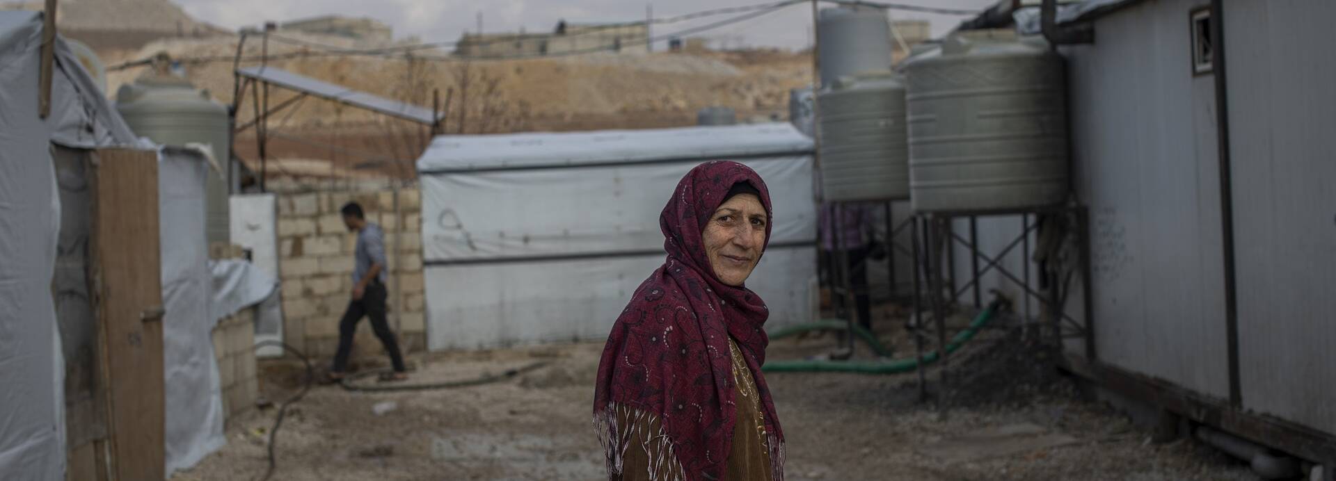 Eine Frau in einem Geflüchtetenlager im Libanon Eine Frau schaut in die Kamera, im Hintergrund ist ein Geflüchtetenlager zu sehen.