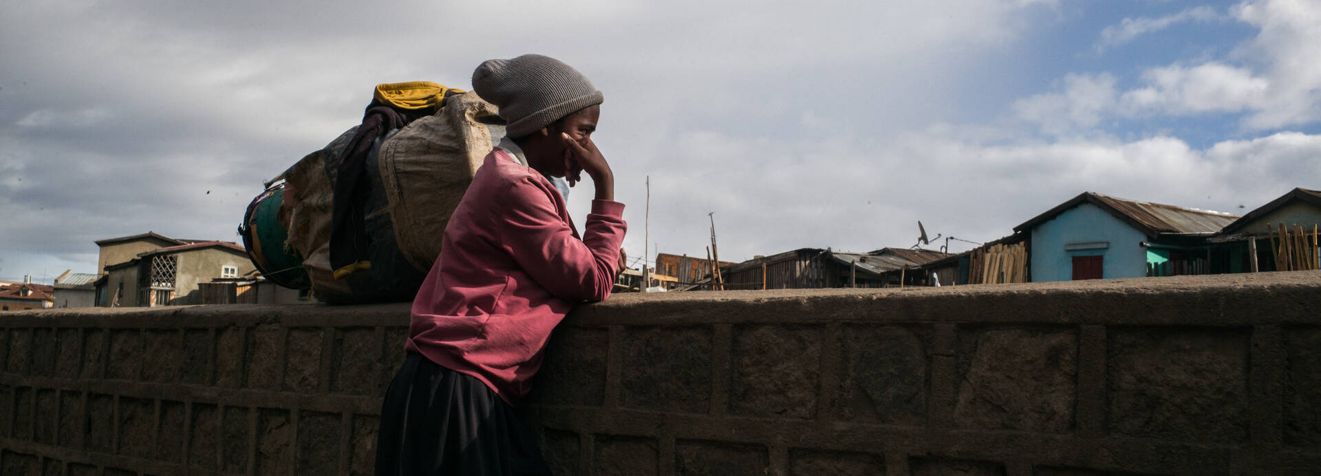 In Madagaskar leben 80,3 Prozent der Bevölkerung in extremer Armut. Eine jugendliche Person steht vor einer Mauer, den Kopf auf den rechten Arm gestützt und blickt gedankenverloren auf die Wohnhäuser des Arbeiter*innenviertels Antohamadinik. Es gilt als eines der prekärsten und ungesündesten in der Stadt Antananarivo in Madagaskar.