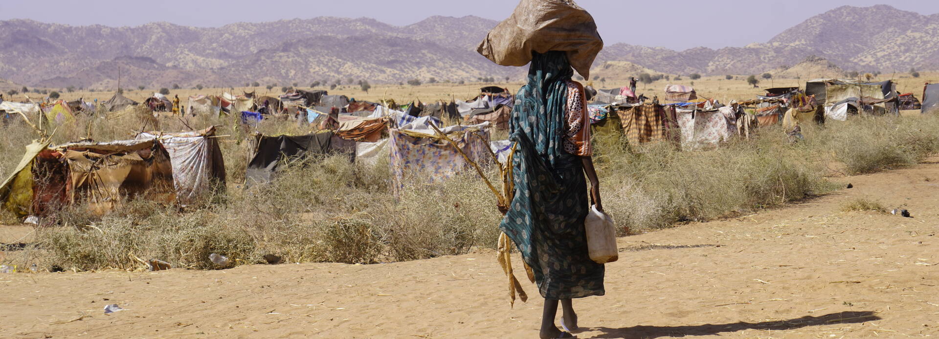 Eine Person im Sudan mit Gepäck auf dem Kopf geht durch eine trockene Landschaft, im Hintergrund ist ein provisorisches Lager zu sehen Eine Person im Sudan mit Gepäck auf dem Kopf geht durch eine trockene Landschaft, im Hintergrund ist ein provisorisches Lager zu sehen