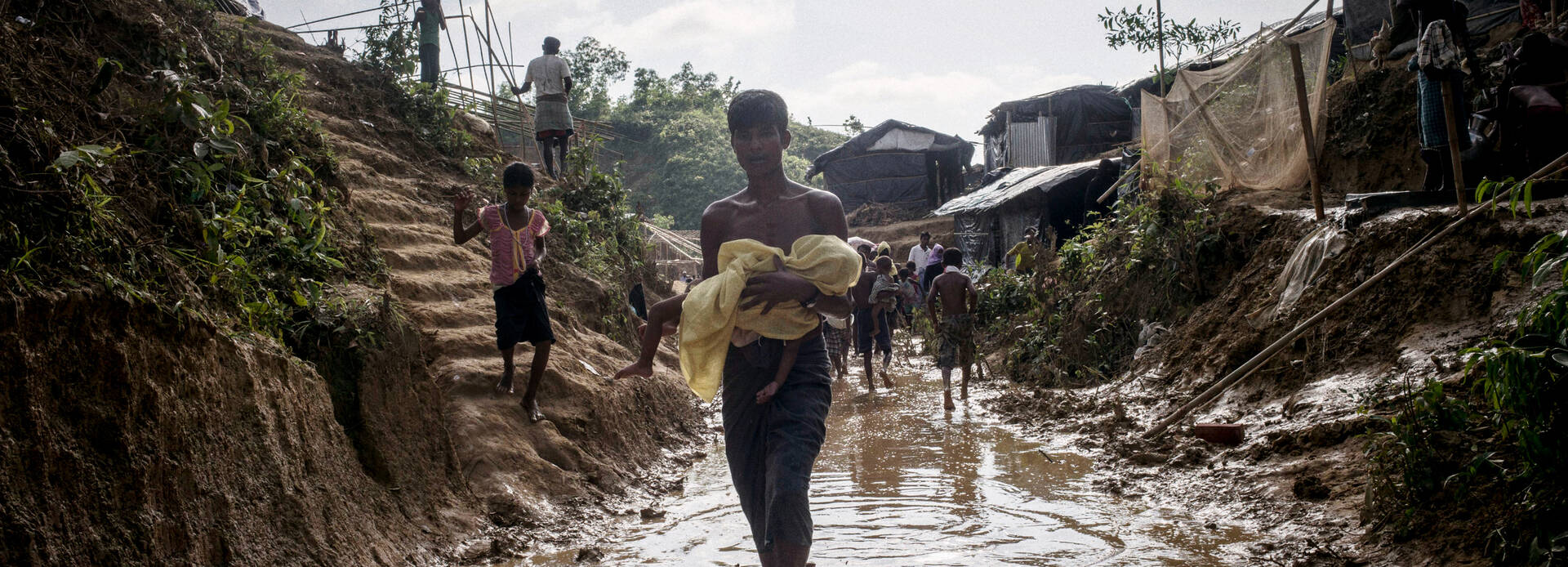 Junger Rohingya trägt in ein Tuch gewickeltes Baby durch knöcheltiefes Wasser in einem Flüchtlingscamp in Bangladesch