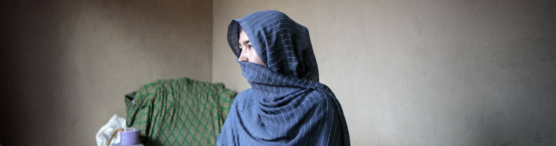 Eine Frau aus Afghanistan sitzt in ihrem Zuhause und schaut zur Seite. Ein blaues Tuch bedeckt ihren Kopf und ihren Mund.