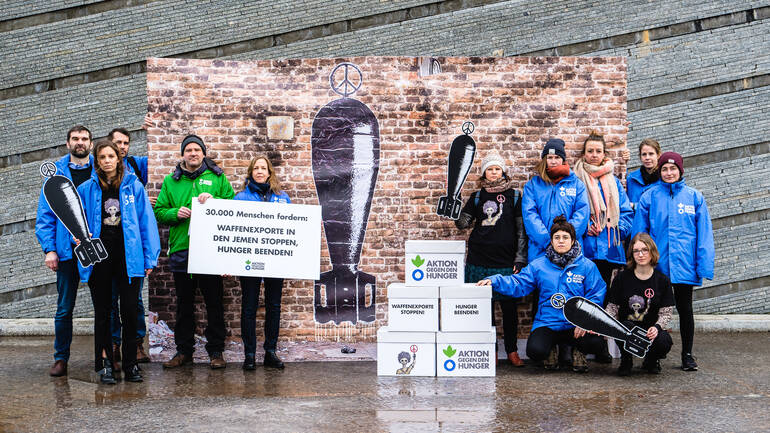 Team von Aktion gegen den Hunger zusammen mit den Unterschriften-Boxen vor einem Banner des jemenitischen Künstlers Murad Subay.