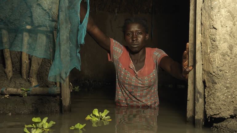 Eine Frau steht in der Tür ihres Hauses mitten im Wasser, vor ihr schwimmen Pflanzen,