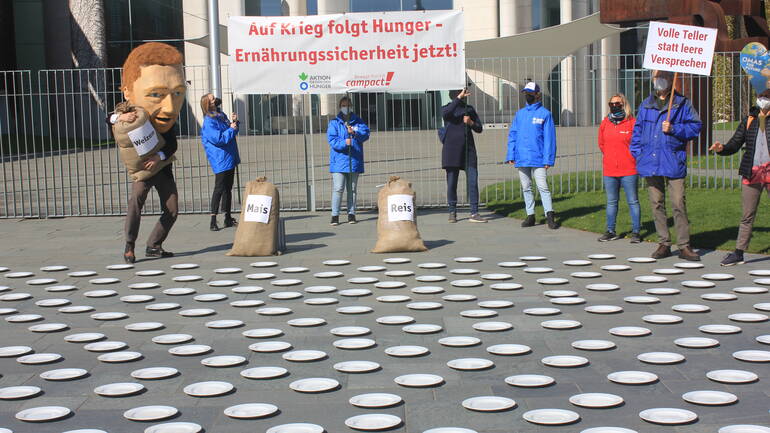 Christian Lindner, Aktivist*innen von Aktion gegen den Hunger und Campact und Demonstrierende stehen um Säcke mit Weizen, Reis und Mais vor 282 Tellern.