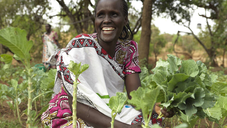 Rosina aus Kenia sitzt in ihrem Gemeinschaftsgarten, pflückt Gemüse und lacht.