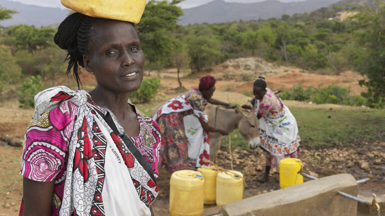 Rosina steht mit einem gelben Wasserkanister auf dem Kopf vor dem Bohrloch, im Hintergrund zwei Frauen mit einem Esel und weiteren Wasserkanistern.