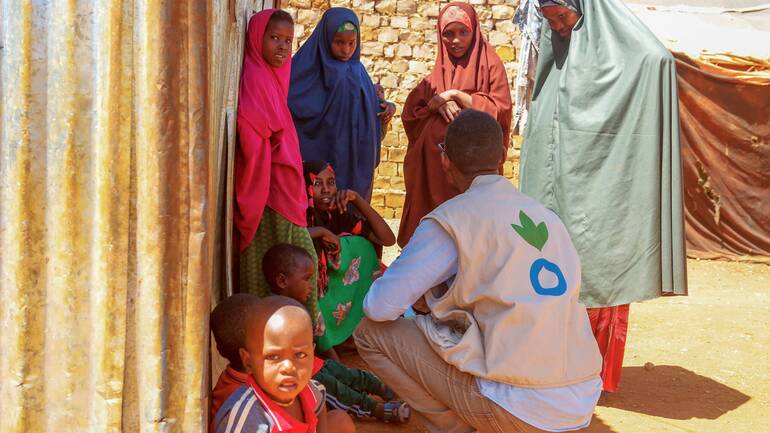 Ein Mitarbeiter von Aktion gegen den Hunger ist im Gespräch mit Frauen und Kindern aus Somalia – alle suchen Schatten hinter einer Wellblechwand.