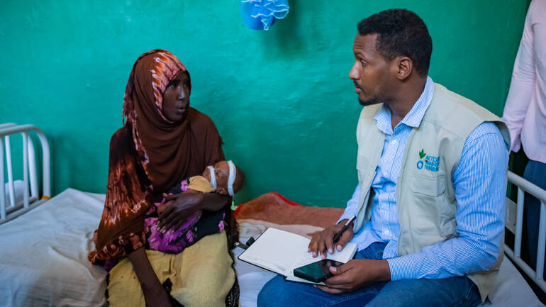 Eine Mutter sitzt mit ihrem mangelernährten Kind auf dem Arm auf dem Bett eines Krankenhauses in Somalia und spricht mit einem Mitarbeiter von Aktion gegen den Hunger