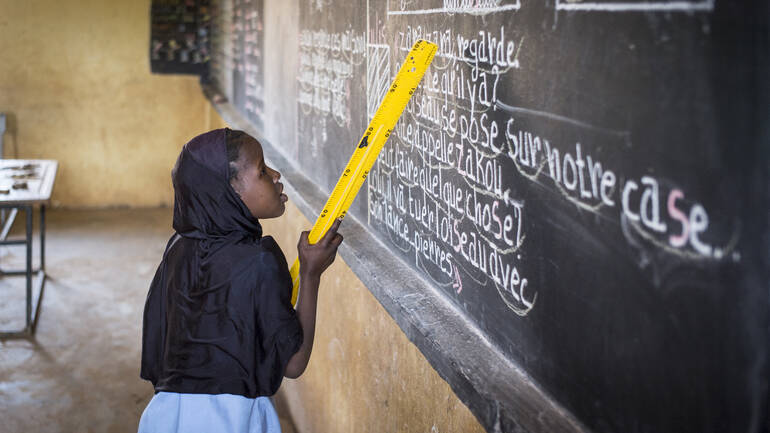 Eine Schülerin zeigt mithilfe eines gelben Lineals auf einen Text auf einer Tafel in einer Schule