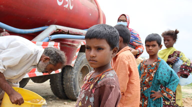 Im Hintergrund ein Wasser-Tanklaster, ein Mann kippt sauberes Wasser in einen Kanister, daneben stehen Kinder