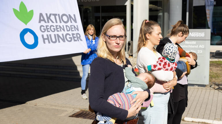 Drei Mütter stehen mit ihren Babys vor der Nestlé-Zentrale in Frankfurt, im Hintergrund hält eine Mitarbeiterin von Aktion gegen den Hunger ein Banner.
