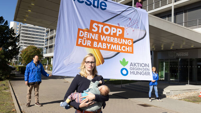 Eine Mutter stillt ihr Baby vor einem Banner mit der Aufschrift "Nestlé, Stopp Deine Werbung für Babymilch!", gehalten von Mitarbeitenden von Aktion gegen den Hunger