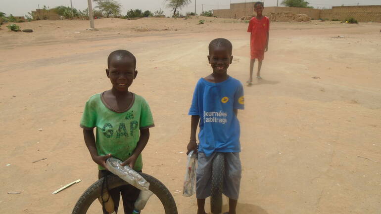 Drei Jungs aus Touba Séras lächeln in die Kamera, zwei haben alte Reifen und Plastikflaschen dabei