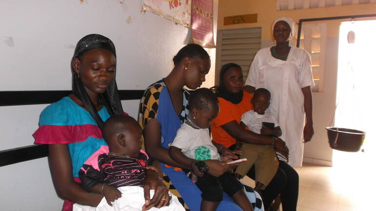 Drei Mütter warten mit ihren Kindern auf dem Schoß auf Behandlung im Gesundheitszentrum, eine Krankenschwester steht im Hintergrund