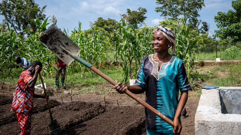 Eine Frau aus der Zentralafrikanischen Republik hält lächelnd ihre Schaufel hoch, während im Hintergrund andere Menschen einen Acker bestellen.