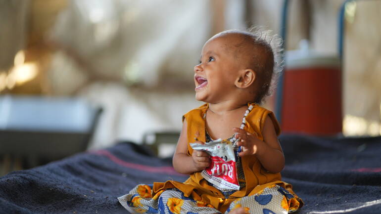 Eines der Kinder von Ali Ahmed im Gesundheitszentrum, lachend mit einer Packung RUTF in der Hand. Eines der Kinder von Ali Ahmed im Gesundheitszentrum, lachend mit einer Packung RUTF in der Hand.
