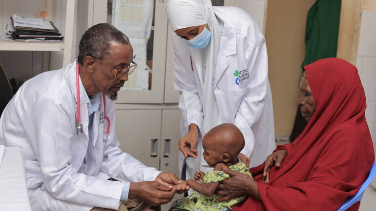 Eine Untersuchung von einem Kind in Somalia - der Arzt hält prüfend und liebevoll die Hand des Mädchens, das auf dem Schoß der mutter sitzt. Die Schwester gibt dem Kind Stabilität. Eine Untersuchung von einem Kind in Somalia - der Arzt hält prüfend und liebevoll die Hand des Mädchens, das auf dem Schoß der mutter sitzt. Die Schwester gibt dem Kind Stabilität.