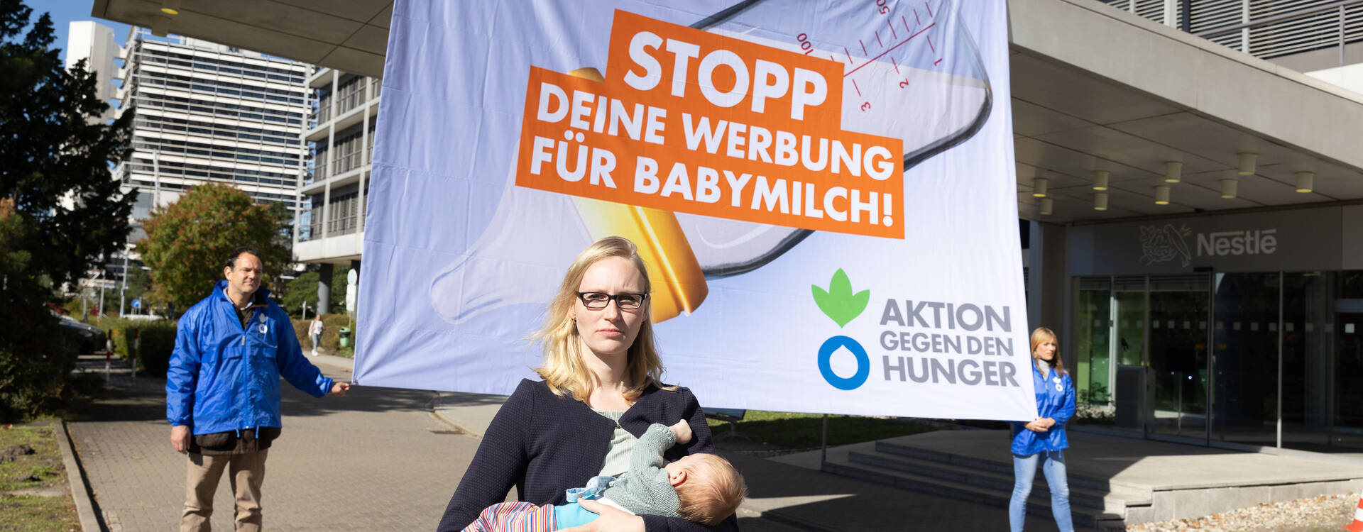 Eine Mutter stillt ihr Baby vor einem Banner mit der Aufschrift "Nestlé, Stopp Deine Werbung für Babymilch!", gehalten von Mitarbeitenden von Aktion gegen den Hunger