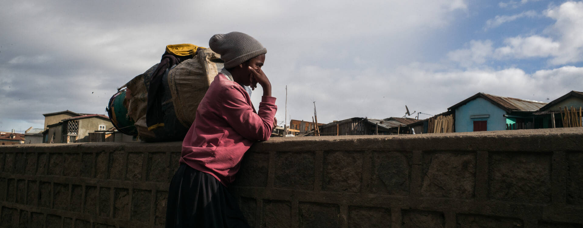 In Madagaskar leben 80,3 Prozent der Bevölkerung in extremer Armut. Eine jugendliche Person steht vor einer Mauer, den Kopf auf den rechten Arm gestützt und blickt gedankenverloren auf die Wohnhäuser des Arbeiter*innenviertels Antohamadinik. Es gilt als eines der prekärsten und ungesündesten in der Stadt Antananarivo in Madagaskar.