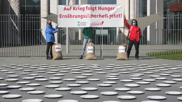 Drei Aktivist*innen von Aktion gegen den Hunger und Campact mit Banner "Auf Krieg folgt Hunger – Ernährungssicherheit jetzt!", mit drei Säcken Mais, Reis und Weizen vor 282 Tellern.