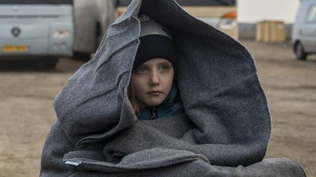 Ein Mädchen sitzt in eine Decke gehüllt neben einem Rollkoffer vor zwei Bussen an der Grenze während der Flucht aus der Ukraine.