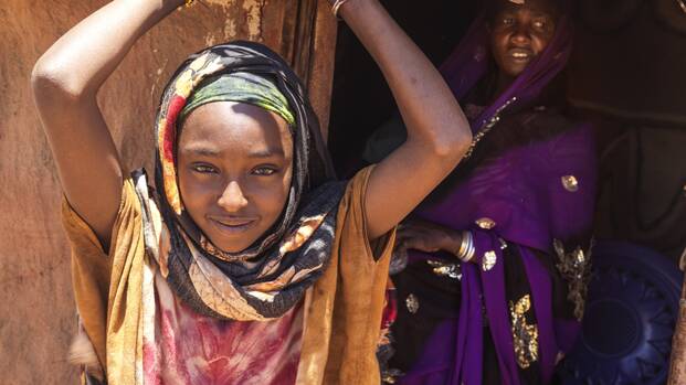Ein junges Mädchen aus Äthiopien in buntem Tuch hebt die Arme und lächelt in die Kamera, im Hintergrund steht eine Frau mit lila Tuch.