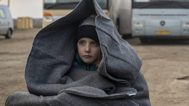 Ein MÃ¤dchen sitzt in eine Decke gehÃ¼llt neben einem Rollkoffer vor zwei Bussen an der Grenze wÃ¤hrend der Flucht aus der Ukraine.
