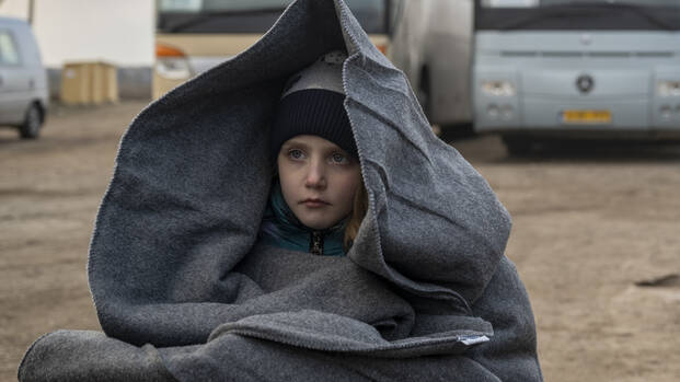 Ein Mädchen sitzt in eine Decke gehüllt neben einem Rollkoffer vor zwei Bussen an der Grenze während der Flucht aus der Ukraine.