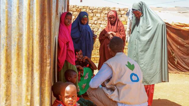 Ein Mitarbeiter von Aktion gegen den Hunger ist im Gespräch mit Frauen und Kindern aus Somalia – alle suchen Schatten hinter einer Wellblechwand.