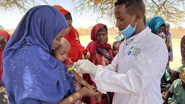 Ein Mitarbeiter von Aktion gegen den Hunger untersucht in Somalia ein Kleinkind auf den Armen seiner Mutter auf Mangelernährung, im Hintergrund warten weitere Mütter mit Kindern.