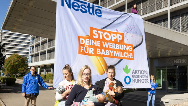 MÃ¼tter mit Babys stillen vor der NestlÃ©-Zentrale in Frankfurt am Main, im Hintergrund halten Aktivist*innen von Aktion gegen den Hunger ein Banner: NestlÃ©: Stopp deine Werbung fÃ¼r Babymilch!