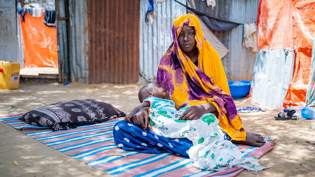 Eine junge Frau aus Somalia sitzt mit ihrem Baby in Tücher gehüllt auf einer Decke in einem Lager und schaut mit leerem Ausdruck zur Seite.