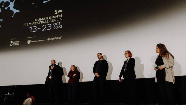 John Shipton, Stella Assange und Gabriel Shipton zusammen mit Anna Ramskogler-Witt und Moderatorin Sham Jaff auf der Bühne, auf der Leinwand das Logo des Human Rights Film Festival Berlin