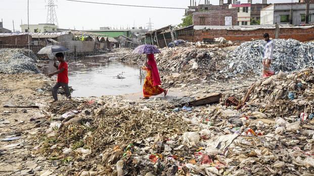 Drei Menschen aus Bangladesch laufen mit Schirmen durch ein Feld voller Abfälle einer Gerberei