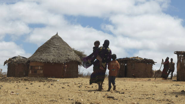 Eine Mutter läuft mit ihren Kindern durch die karge Landschaft Äthiopiens in ihr Dorf, im Hintergrund stehen zwei weitere Personen neben Hütten