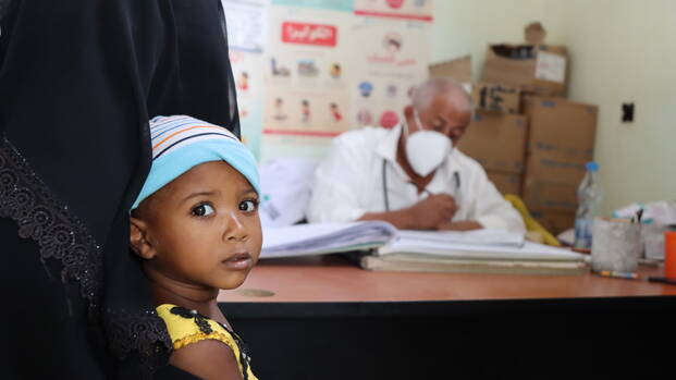 Ein Kind in den Armen der Mutter schaut in die Kamera, im Hintergrund ein Arzt aus dem Gesundheitszentrum in Almakhzan im Jemen.