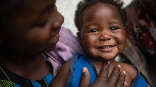 Ein Kleinkind schaut lachend in die Kamera, während die Mutter es beschützend hält und anlächelt