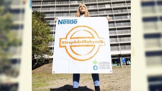 Unsere Kollegin steht vor der Nestlé-Zentrale in Frankfurt am Main und hält ein Banner mit einem orangefarbenen Stempel mit dem Hashtag #StopAds4Babymilk hoch.