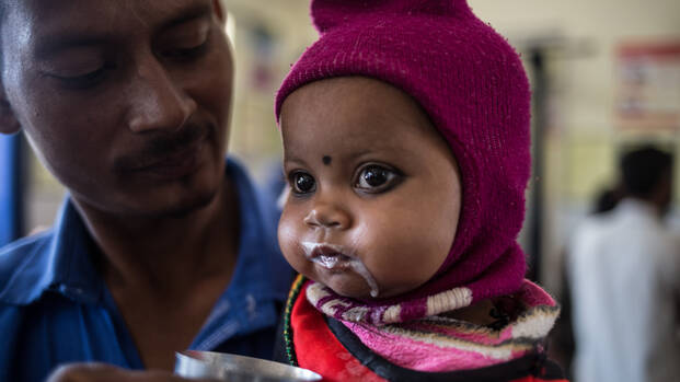 Bhumika bekommt in der Klinik in Indien Aufbaunahrung in einem Becher von ihrem Vater.