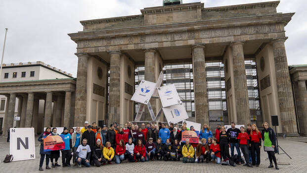 Mitglieder von Hilfsorganisationen bei der Protestaktion #Luftnachoben vor dem Brandenburger Tor in Berlin, im Hintergrund ein zusammenfallendes Kartenhaus.