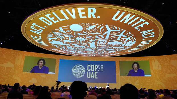 Im Plenarsaal der Klimakonferenz in Dubai sitzen die Vertreter*innen der Staaten und hören der Sprecherin vorne zu. Über allen wölbt sich eine große Kuppel mit der Aufschrift "Unite, Act, Deliver".