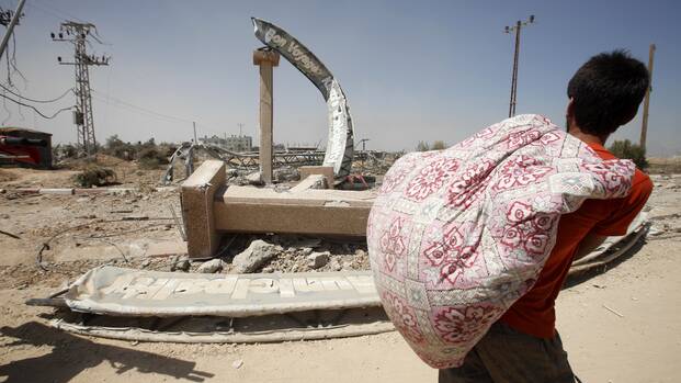 Ein junger Mann trägt einen Sack mit Habseligkeiten auf dem Rücken und schaut auf einen zerstörten Torbogen am Eingang eines zerbombten Ortes.